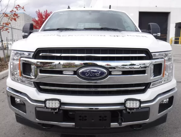 Two of the lamps mounted low on the front bumper of a white Ford pickup lettered CONSERVATION OFFICER